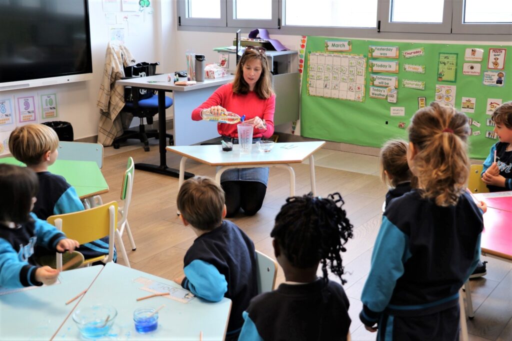 La imagen muestra a niños pequeños observando atentamente un experimento científico llevado a cabo por su profesora, mostrando curiosidad y aprendizaje.